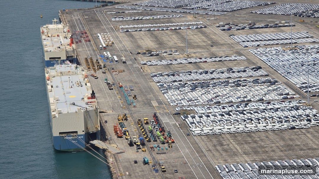 An aerial view of a busy port with several large car carrier ships docked and vast lots filled with rows of parked vehicles. Construction equipment and trucks are also visible on the docks.