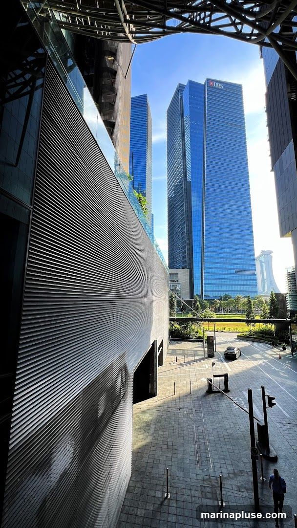 A view looking up at modern skyscrapers with a distinctive ribbed facade in the foreground. The DBS logo is visible on one of the tall blue glass buildings in the background. The scene is set during the day with a clear blue sky and sunlight casting shadows on the paved ground. A car is seen driving on the road, and a person is jogging on the sidewalk.