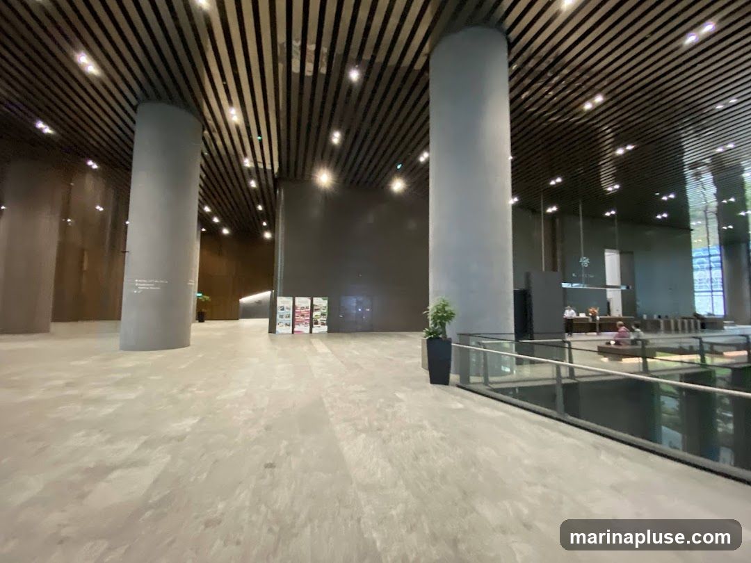 A wide shot of a spacious and modern office lobby with large cylindrical columns and a slatted ceiling. The floor is made of large, light-colored tiles. In the background, there is a reception desk with a few people, and some display boards. A glass railing overlooks a lower level.