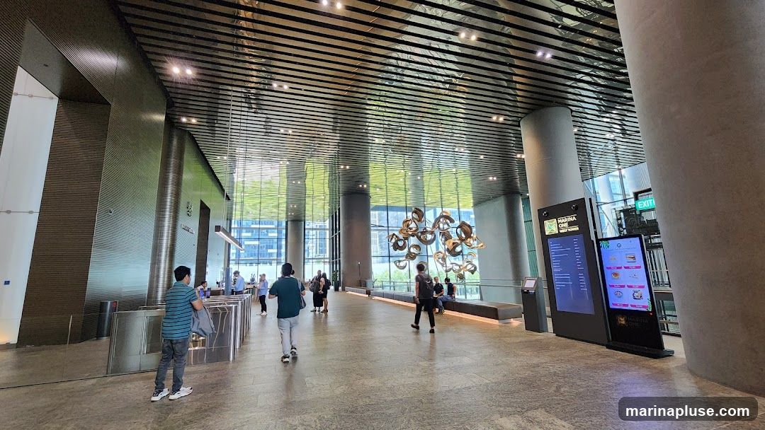 A wide shot of a spacious and modern office building lobby. The ceiling features a reflective, slatted design that mirrors the greenery visible through the large windows. A contemporary art sculpture hangs in the center of the space. Several people are seen walking through the lobby, and a digital display board shows information and images, including what appears to be food items.