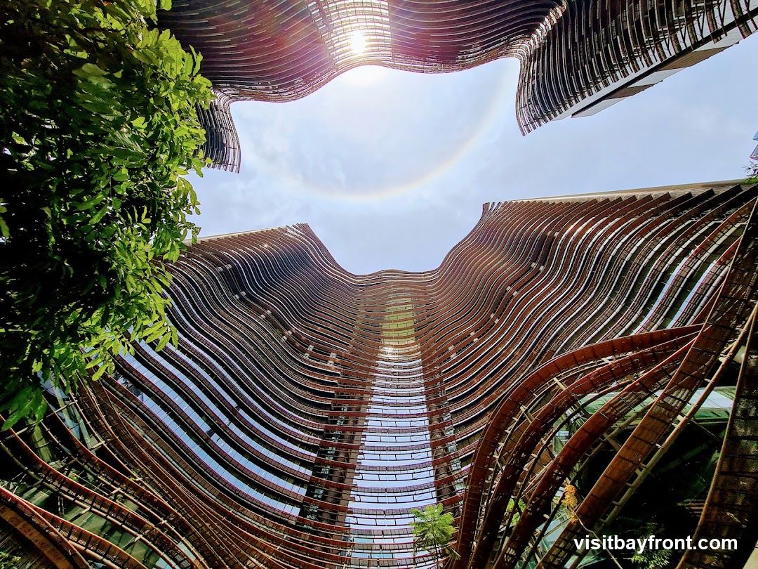 A low-angle shot looking up at a modern building with a unique wavy facade. The exterior is covered in horizontal, curved elements that create a sense of movement. Green foliage is visible on the left side, and the sky above shows a sun halo.