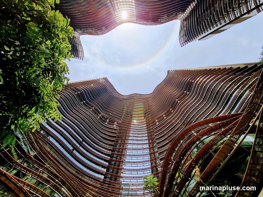A low-angle shot looking up at a modern building with a unique wavy facade. The exterior is covered in horizontal, curved elements that create a sense of movement. Green foliage is visible on the left side, and the sky above shows a sun halo.