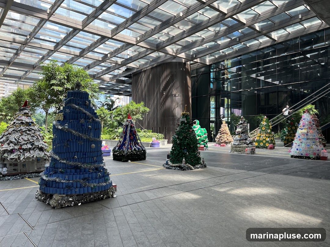 A collection of creatively decorated Christmas trees are displayed in an outdoor plaza with a modern glass-covered ceiling. The trees are made from various recycled materials, including newspapers, blue tubes, and plastic bottles. Some trees are adorned with tinsel, ornaments, and Santa hats. In the background, a person is visible on a staircase inside a building.