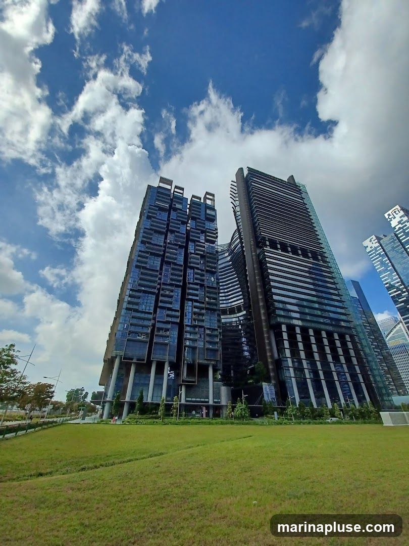 A wide-angle shot captures several modern skyscrapers with distinctive architectural designs against a bright blue sky filled with fluffy white clouds. In the foreground, a lush green lawn slopes gently, creating a park-like setting. The buildings feature glass facades and intricate structural elements, reflecting the daylight. One building on the right has a visible 'Citi' logo.