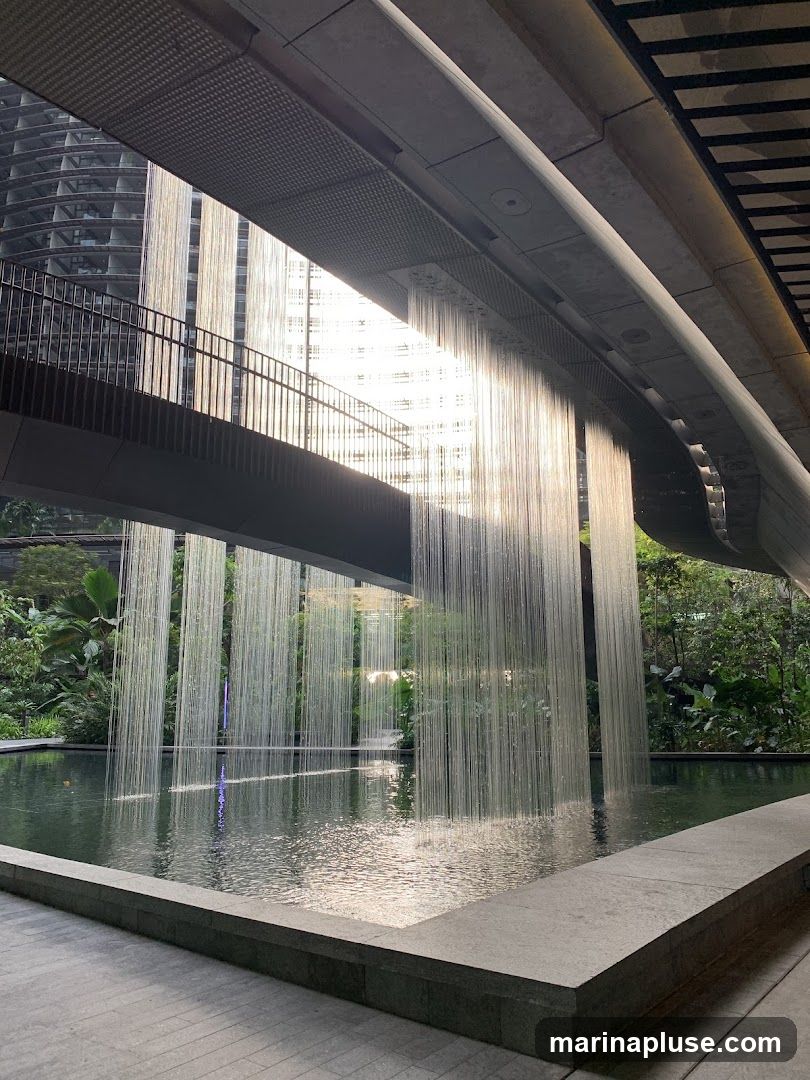 A contemporary architectural design featuring a man-made waterfall cascading into a pool. The water falls in thin, shimmering strands, creating a curtain-like effect. Above the waterfall, a walkway or bridge is visible, with modern buildings in the background. Lush greenery surrounds the water feature, suggesting an urban oasis or a landscaped public space. The lighting creates a serene and reflective atmosphere.