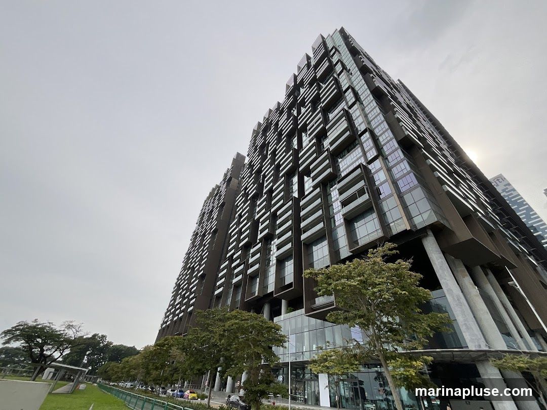 A low-angle shot of a modern, multi-story building with a distinctive facade featuring numerous balconies and glass panels. The building has a dark, textured exterior and is supported by large concrete pillars at its base. Trees and a street with cars are visible in the foreground, under an overcast sky.
