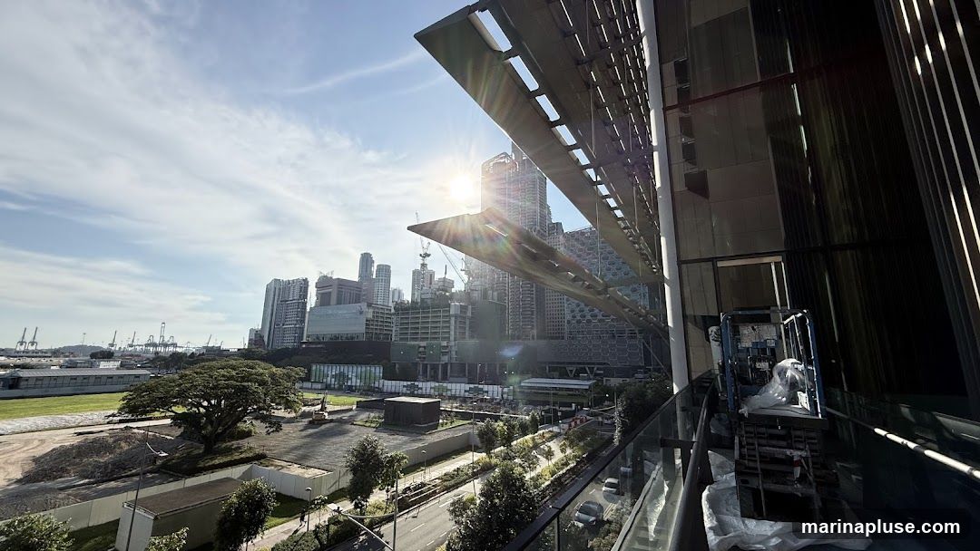 A wide-angle view of a city skyline under a bright, sunny sky with wispy clouds. In the foreground, a modern building with a distinctive angled overhang is visible on the right. Below, a road with some cars can be seen, bordered by greenery and a construction site with heavy machinery and piles of debris. In the distance, a large port with numerous cranes is situated along the coastline, with more city buildings and construction cranes dotting the horizon. The sun is shining brightly, creating lens flare.