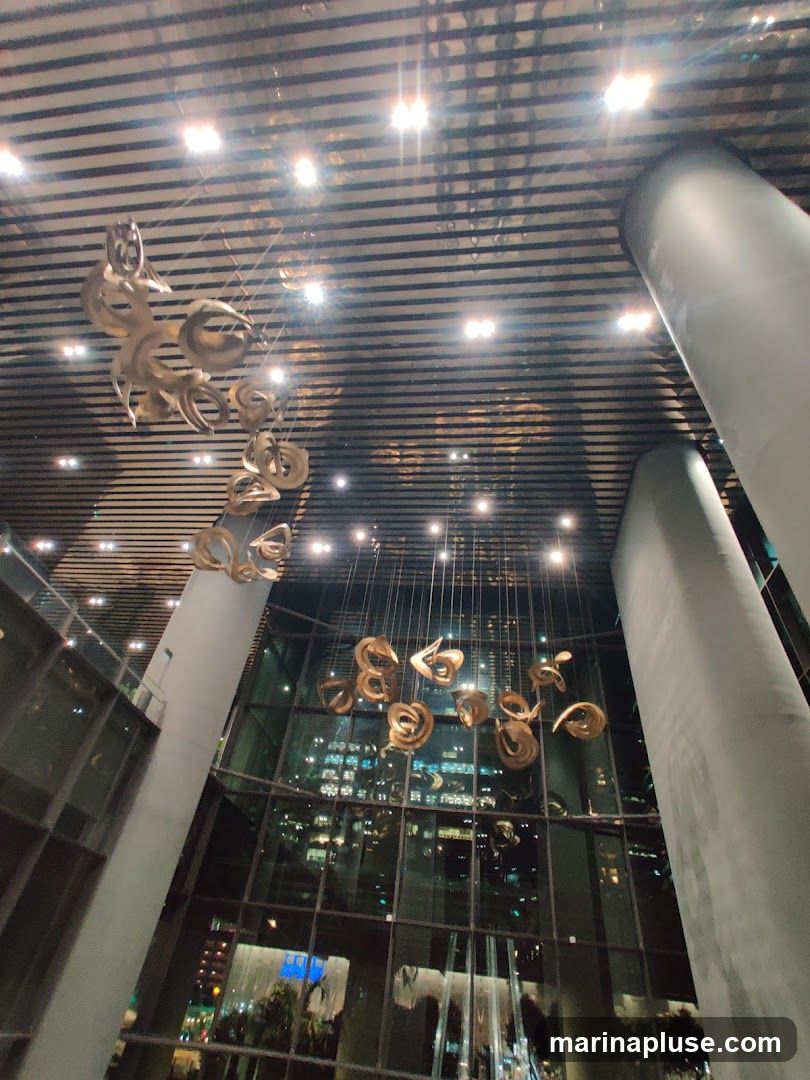 A wide-angle shot of a modern interior space, looking upwards. The ceiling features a striped design with recessed lighting. Several large, abstract, golden sculptures hang from the ceiling by thin wires, creating a dramatic visual effect. Large pillars are visible on the right and left sides of the frame. The background consists of a floor-to-ceiling glass facade, reflecting the lights of a city skyline at night.