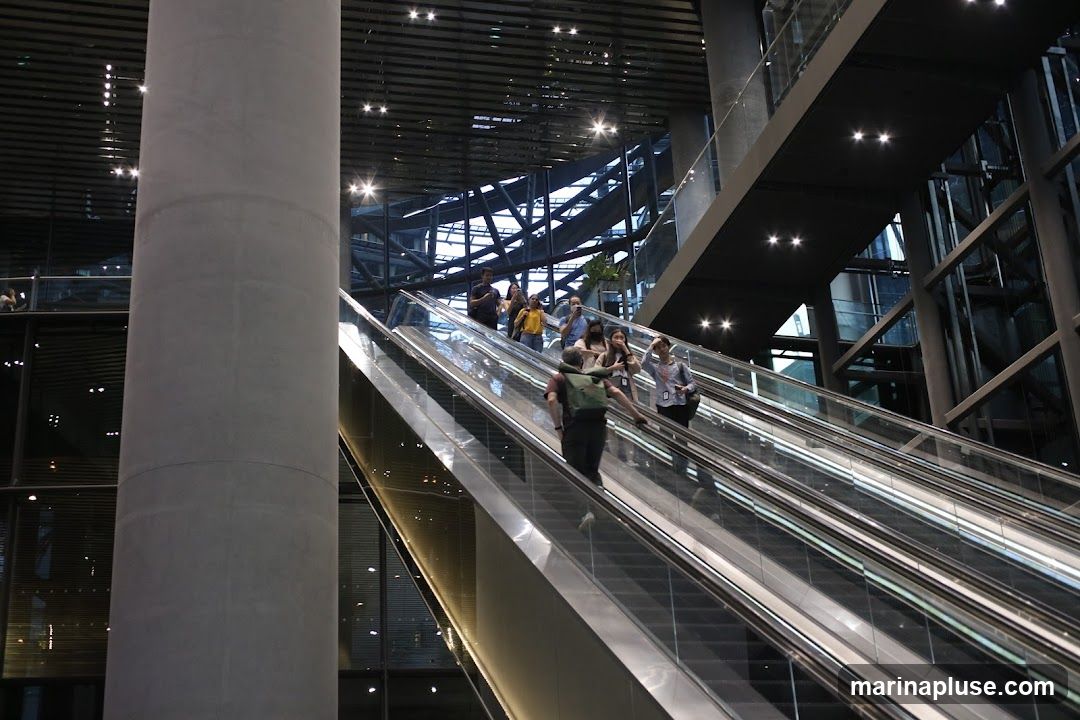 A group of people are riding escalators in a large, modern building with a concrete pillar and glass facade. The escalators are moving upwards and downwards, and several individuals are visible on them. The architecture features a sleek, contemporary design with metal accents and overhead lighting.