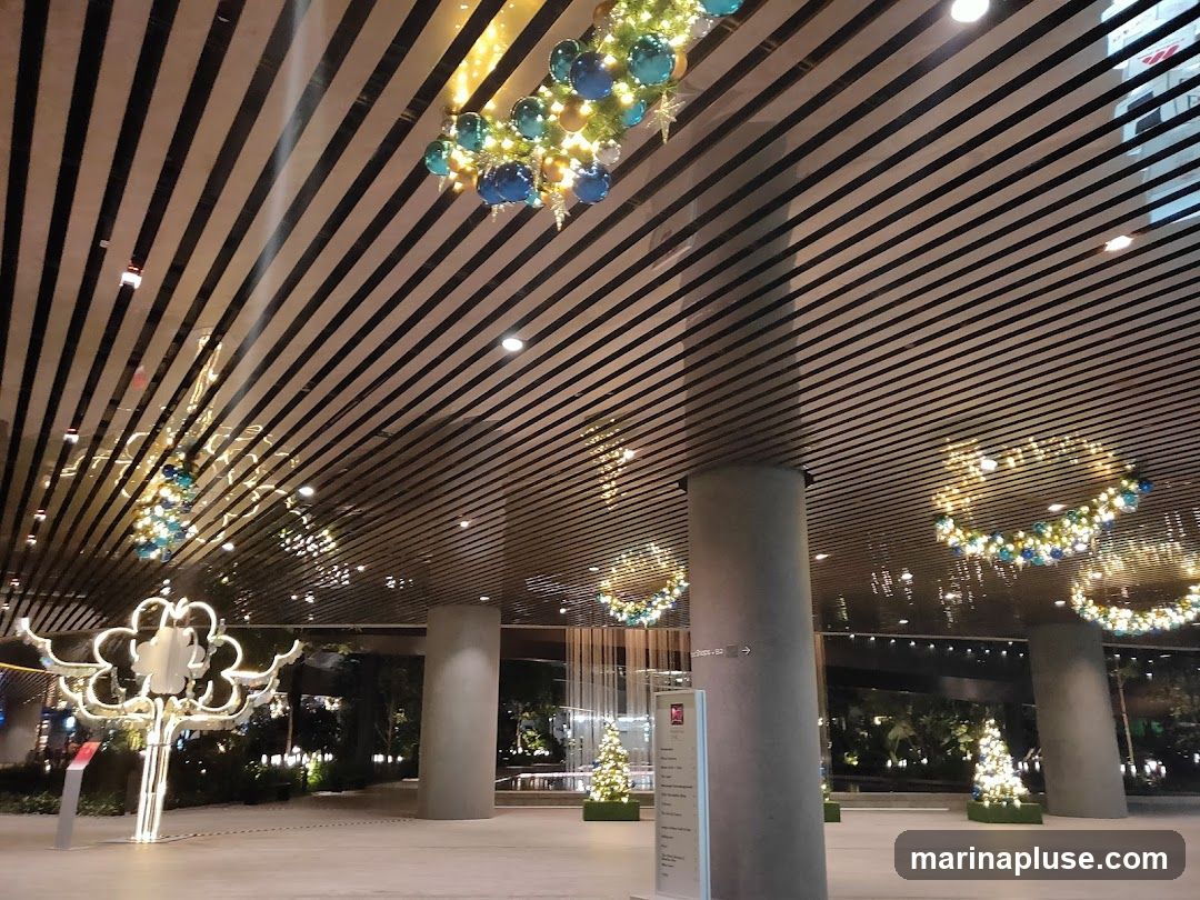 The image shows a modern building interior with a slatted ceiling and large pillars. Festive Christmas decorations are visible, including illuminated wreaths with blue and gold ornaments hanging from the ceiling, and small decorated Christmas trees placed on the floor. A large, illuminated, cloud-shaped sculpture is also present on the left side of the image. The overall ambiance suggests a holiday-themed event or display within a commercial or public space.