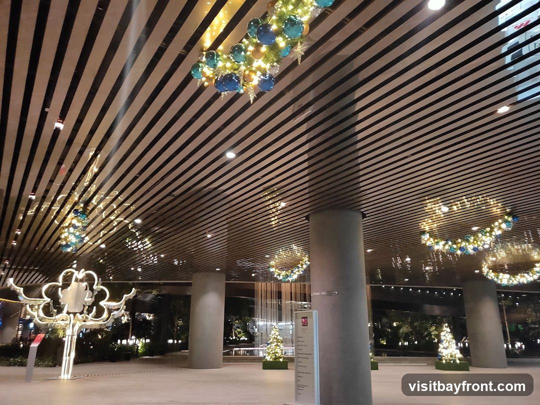 The image shows a modern building interior with a slatted ceiling and large pillars. Festive Christmas decorations are visible, including illuminated wreaths with blue and gold ornaments hanging from the ceiling, and small decorated Christmas trees placed on the floor. A large, illuminated, cloud-shaped sculpture is also present on the left side of the image. The overall ambiance suggests a holiday-themed event or display within a commercial or public space.