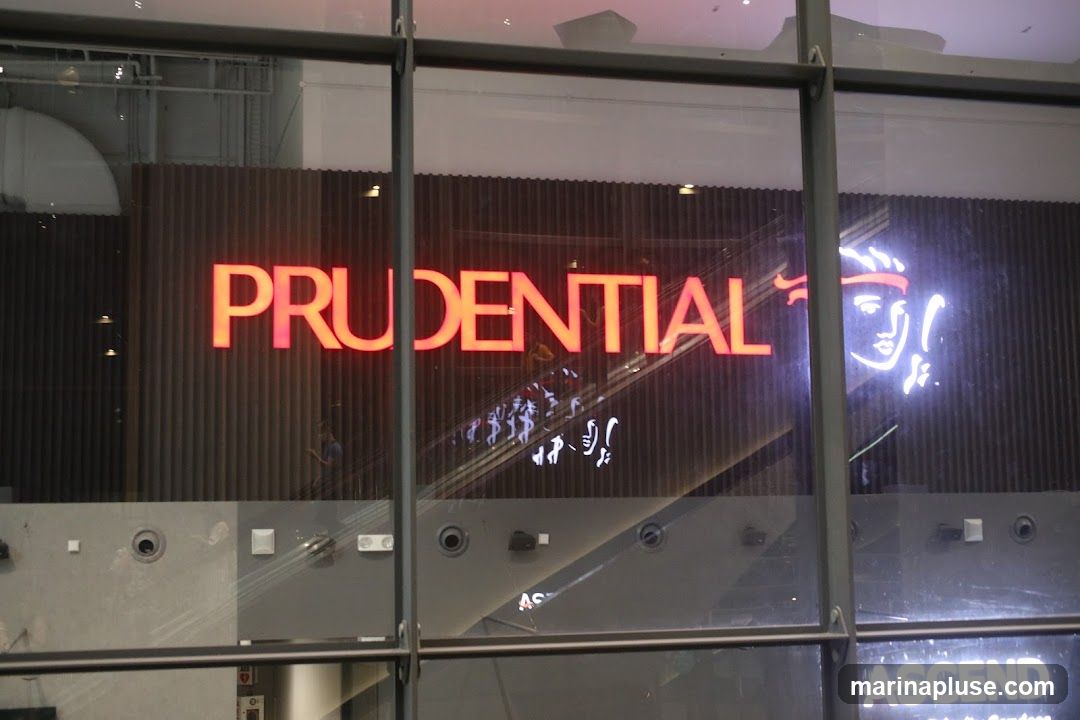A large, illuminated sign with the word "PRUDENTIAL" in orange neon letters is displayed on a dark, ribbed wall. To the right of the sign, a white neon outline of a face with a red headband is visible. Reflections of people and escalators can be seen on the glass in front of the sign.