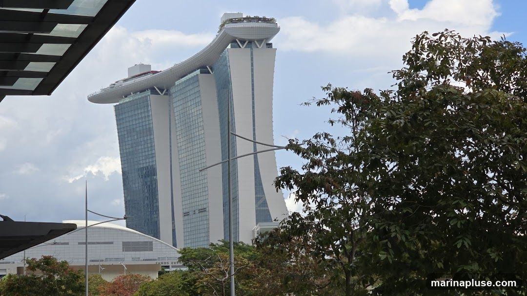 A view of the Marina Bay Sands hotel in Singapore, with its distinctive boat-shaped rooftop, against a cloudy blue sky. In the foreground, a portion of a modern building with a glass roof is visible on the left, and lush green trees partially obscure the view of the hotel on the right. Below the hotel, a large, curved white building with the text "SANDS EXPO" is visible.