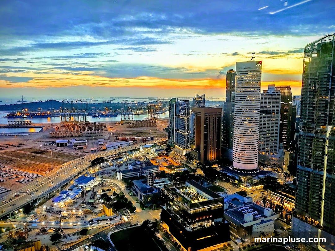 An aerial view of a bustling cityscape at dusk, with a large port in the background. The sky is a vibrant mix of orange, yellow, and blue hues. Tall skyscrapers dominate the foreground, illuminated by interior lights. Below, a highway is visible with car lights, and construction sites with cranes are scattered throughout the urban landscape. The port features numerous shipping containers and cranes, indicating significant industrial activity.