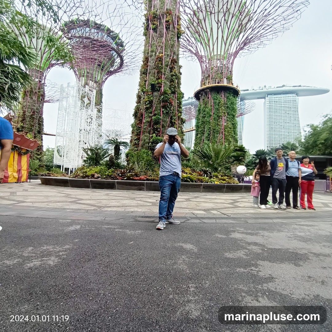 A man stands in front of the Supertree Grove at Gardens by the Bay, Singapore, with a family posing in the background. The iconic Marina Bay Sands hotel is visible in the distance.
