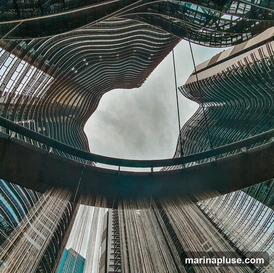 A low-angle shot looking up at the unique, wavy facade of modern buildings, with a circular walkway or structure in the foreground and hanging vines or cables creating a curtain-like effect. The sky is overcast.