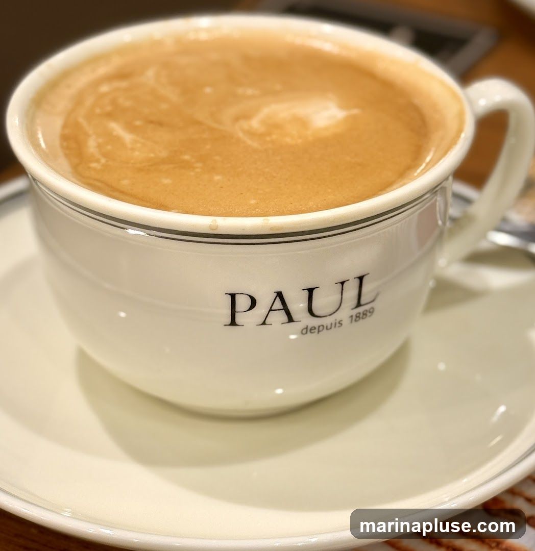 A close-up shot of a white coffee cup with the PAUL logo and "depuis 1889" printed on it. The cup is filled with a frothy coffee drink and sits on a matching saucer.
