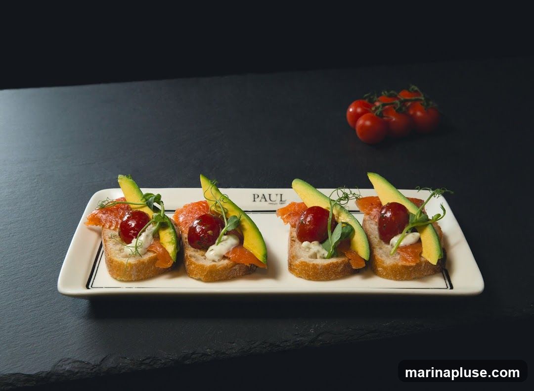 Four small open-faced sandwiches on a white rectangular plate. Each sandwich has a slice of bread topped with smoked salmon, avocado slices, a cherry tomato, and a dollop of white sauce, garnished with microgreens. A small cluster of cherry tomatoes is visible in the background. The plate has the word "PAUL" printed on it.