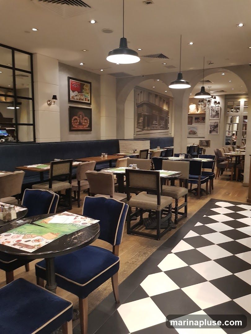 The image shows the interior of a restaurant with a black and white checkered floor. There are several tables and chairs, some with blue velvet upholstery. The walls are decorated with framed pictures and a large mural. Pendant lights hang from the ceiling.
