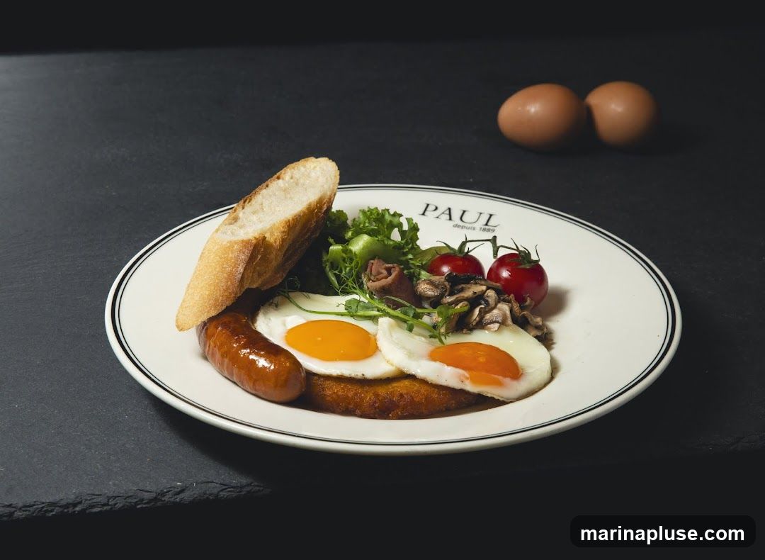 A breakfast plate with two fried eggs, a sausage, a slice of bread, cherry tomatoes, mushrooms, and a small salad. The plate has the PAUL logo and "depuis 1889" printed on it. Two eggs are visible in the background.
