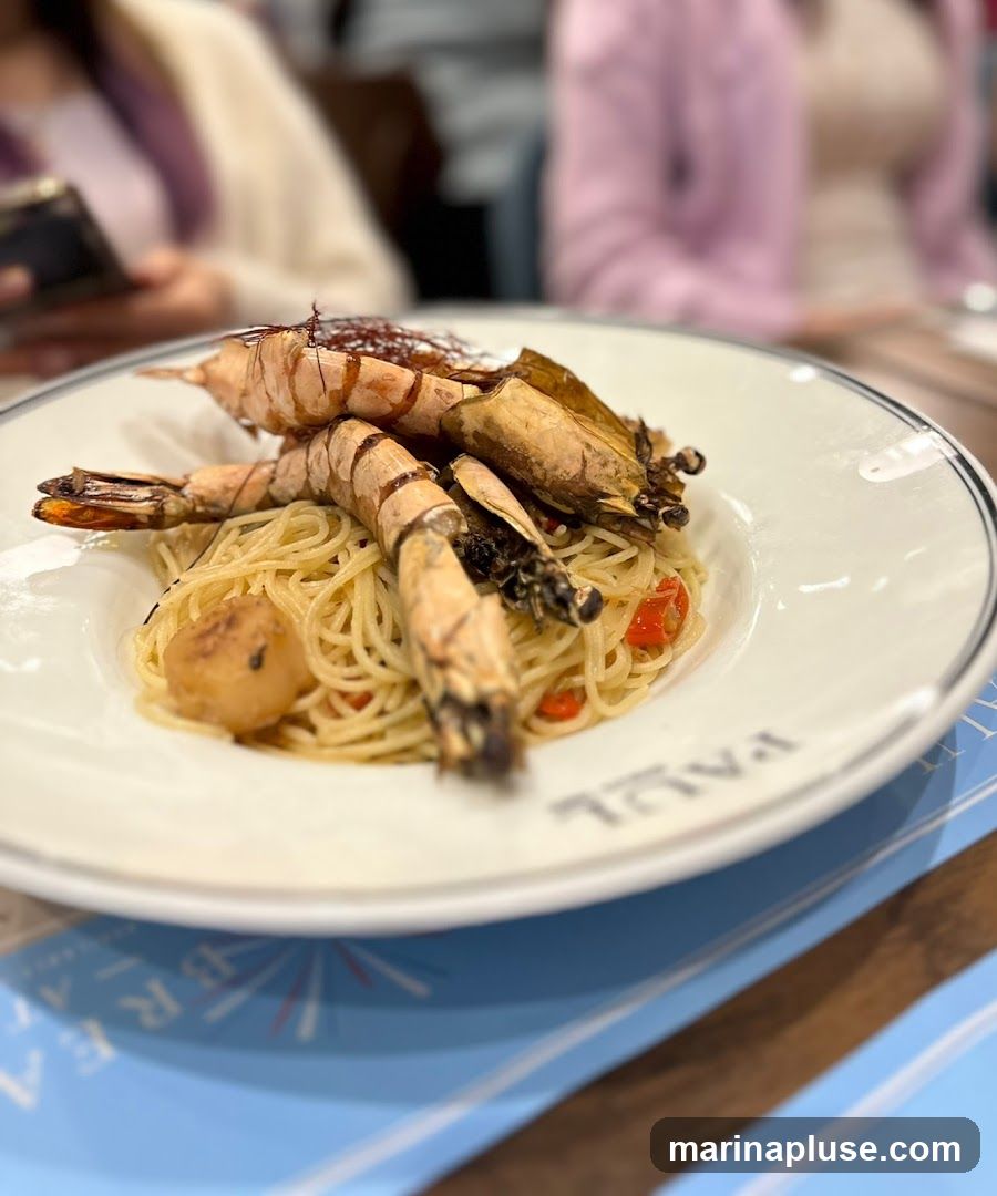 A close-up shot of a plate of spaghetti topped with several large grilled prawns and what appears to be a scallop. The dish is served on a white plate with a black rim, bearing the embossed name "PAUL". In the background, two people are out of focus, one looking at a phone and the other wearing a pink sweater. A blue and white striped placemat is visible in the foreground.