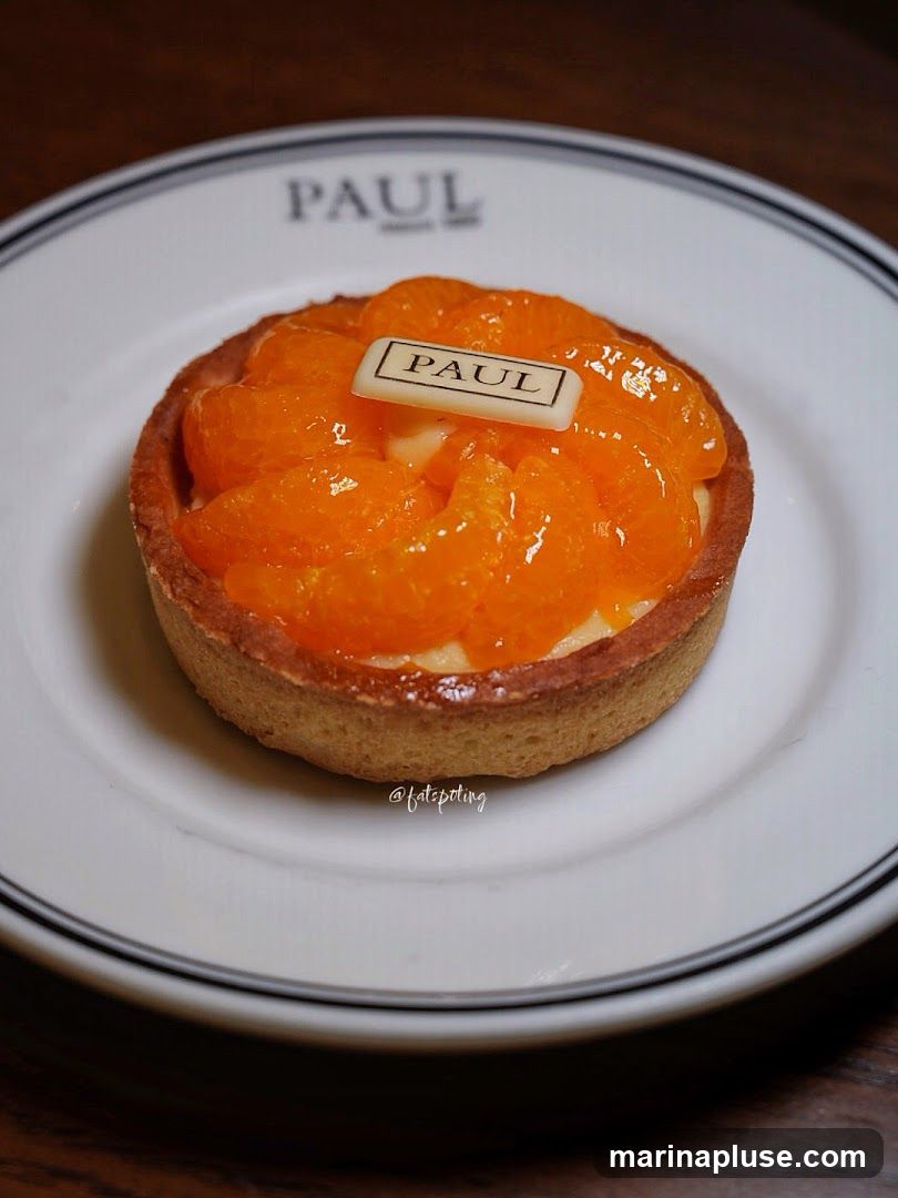 A close-up shot of an orange tart on a white plate with a black rim. The tart has a golden crust and is filled with cream and topped with fresh orange slices. A small white chocolate plaque with the brand name "PAUL" is placed on top of the tart. The plate also has the brand name "PAUL" printed on it.