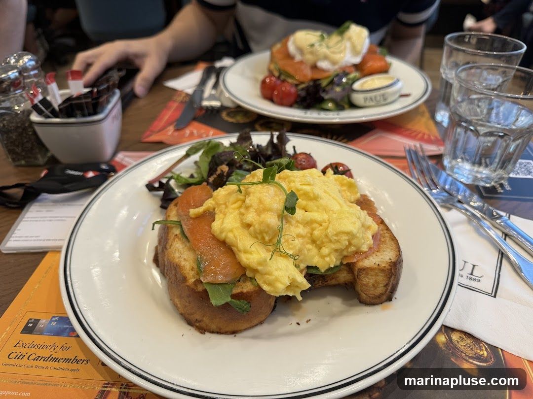 A close-up view of two plates of brunch food, likely at a PAUL bakery. The plate in the foreground features scrambled eggs and smoked salmon on toast, served with a side salad and cherry tomatoes. The plate in the background appears to be Eggs Benedict with smoked salmon. Cutlery, glasses of water, and salt and pepper shakers are also visible on the table. A promotional flyer for Citi Cardmembers is on the table.