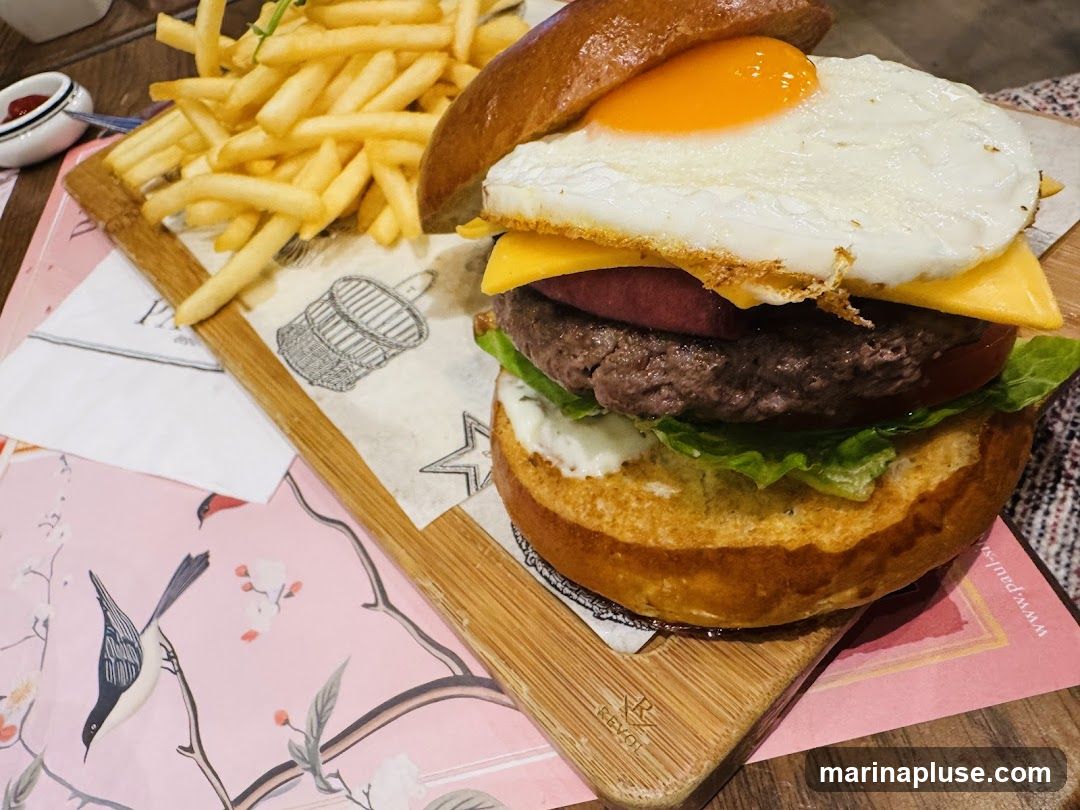 A close-up shot of a gourmet burger served with a side of french fries. The burger is stacked high with a beef patty, cheese, tomato, lettuce, and a fried egg, all nestled within a toasted brioche bun. The fries are golden brown and appear crispy. The meal is presented on a wooden board with decorative paper underneath. A small dish of ketchup is visible in the upper left corner. The table setting includes a pink placemat with a bird and floral motif.