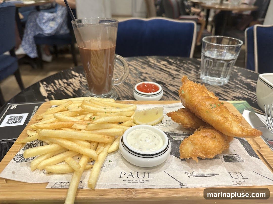 A plate of fish and chips served with tartar sauce, ketchup, and a lemon wedge. A glass of chocolate drink and a glass of water are also on the table. The plate is on a wooden board with a paper liner that has the PAUL logo and text.