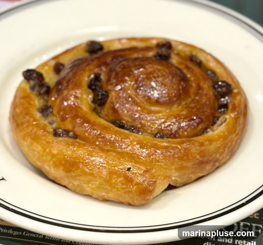 A close-up shot of a golden-brown raisin swirl pastry on a white plate with a black rim. The pastry is glazed and has visible raisins throughout its spiral layers.