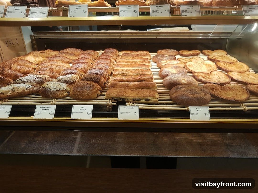 A display case filled with various pastries, including croissants, pain au chocolat, and palmiers. Small labels with names and prices are visible in front of some items. A paper bag with the PAUL logo is also visible.