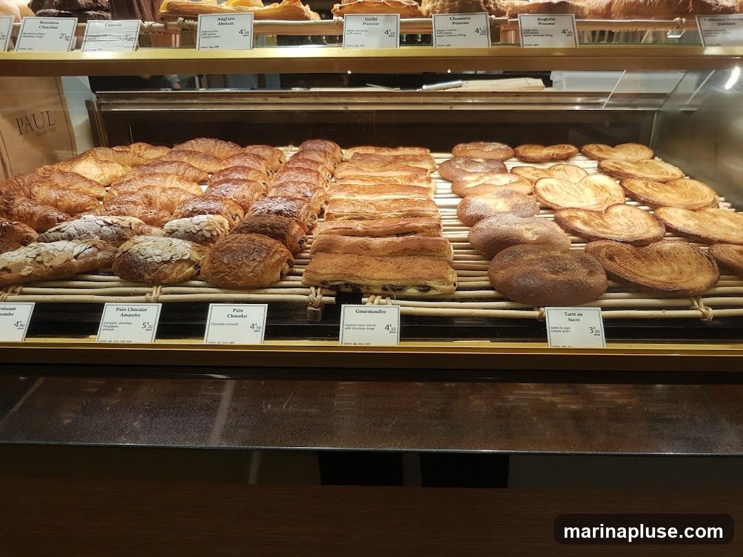 A display case filled with various pastries, including croissants, pain au chocolat, and palmiers. Small labels with names and prices are visible in front of some items. A paper bag with the PAUL logo is also visible.