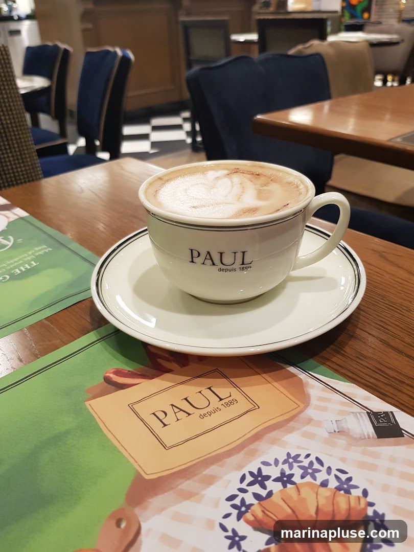 A close-up shot of a cappuccino in a white cup and saucer, with the PAUL logo visible on the cup. The cup sits on a wooden table, with placemats featuring PAUL branding and illustrations of pastries. In the background, blurred seating and a checkered floor suggest the interior of a cafe or restaurant.