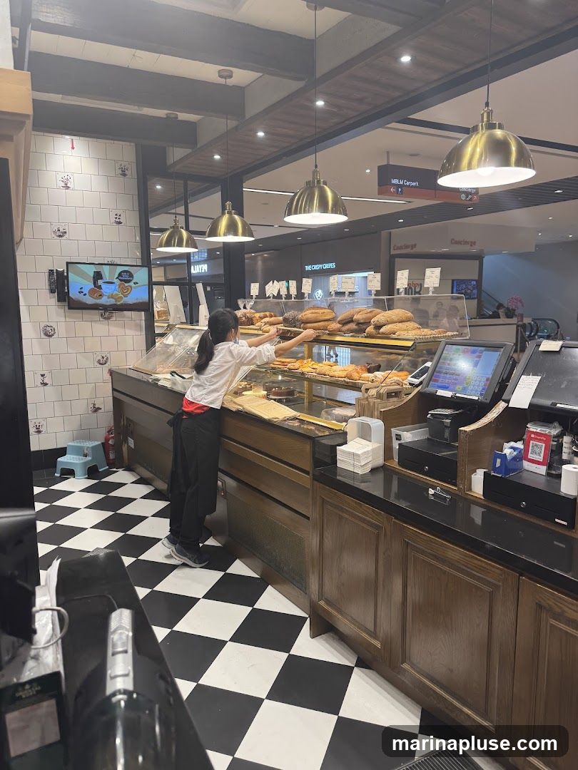 A bakery employee arranges bread and pastries in a display case. The bakery has a black and white checkered floor and wooden cabinets. In the background, there are signs for "The Crispy Crepes" and "Concierge."