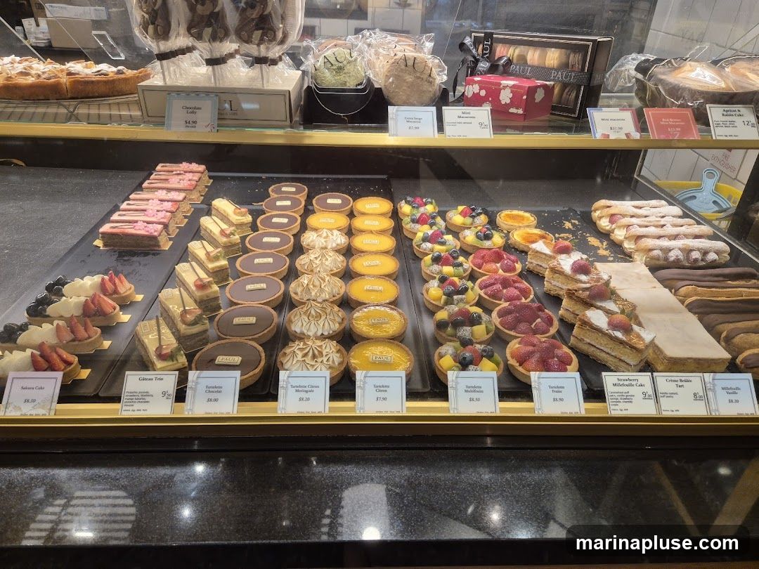 A display case filled with various pastries, tarts, and cakes. In the foreground, there are rows of fruit tarts, chocolate tarts, and lemon meringue tarts. Behind these, there are layered cakes and eclairs. The top shelf shows larger cakes, macarons, and chocolate lollipops.