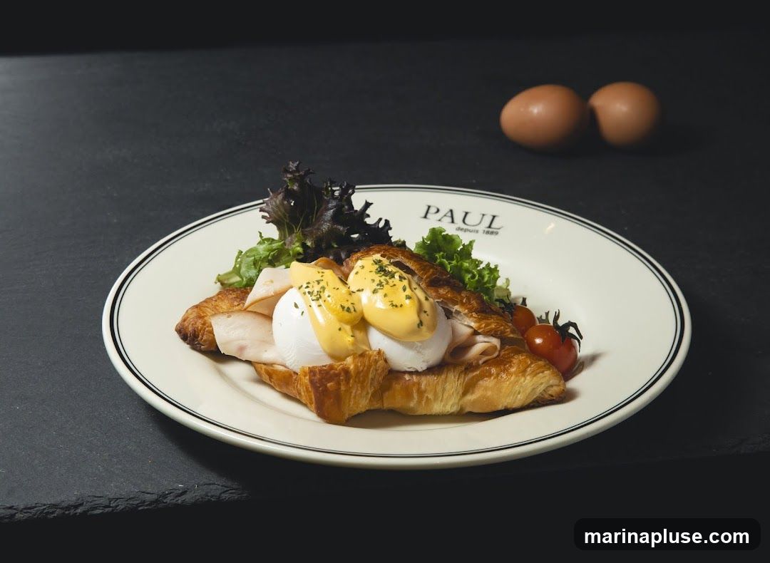 A croissant topped with sliced turkey, poached eggs, hollandaise sauce, lettuce, and cherry tomatoes. The plate has the PAUL logo and "depuis 1889" printed on it. Two eggs are visible in the background.
