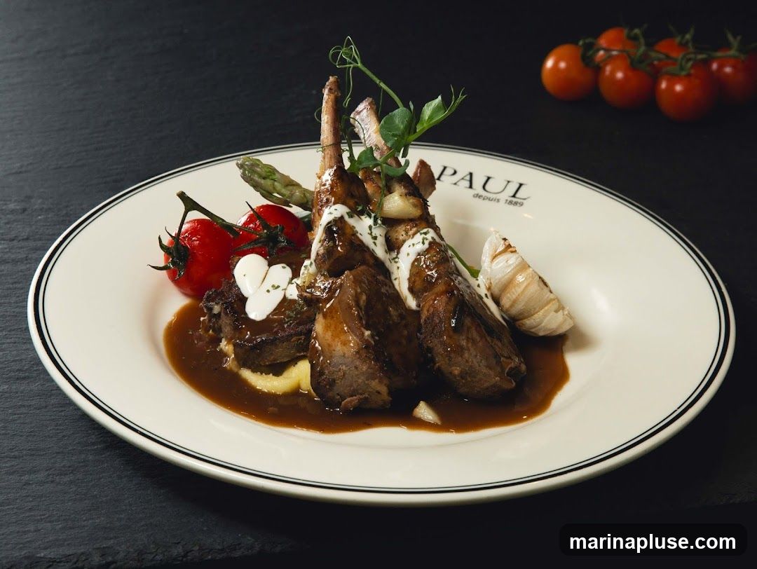 A plate of rack of lamb served with mashed potatoes, asparagus, cherry tomatoes, garlic, and a creamy sauce. The plate has the PAUL logo and the text "depuis 1889" printed on it.