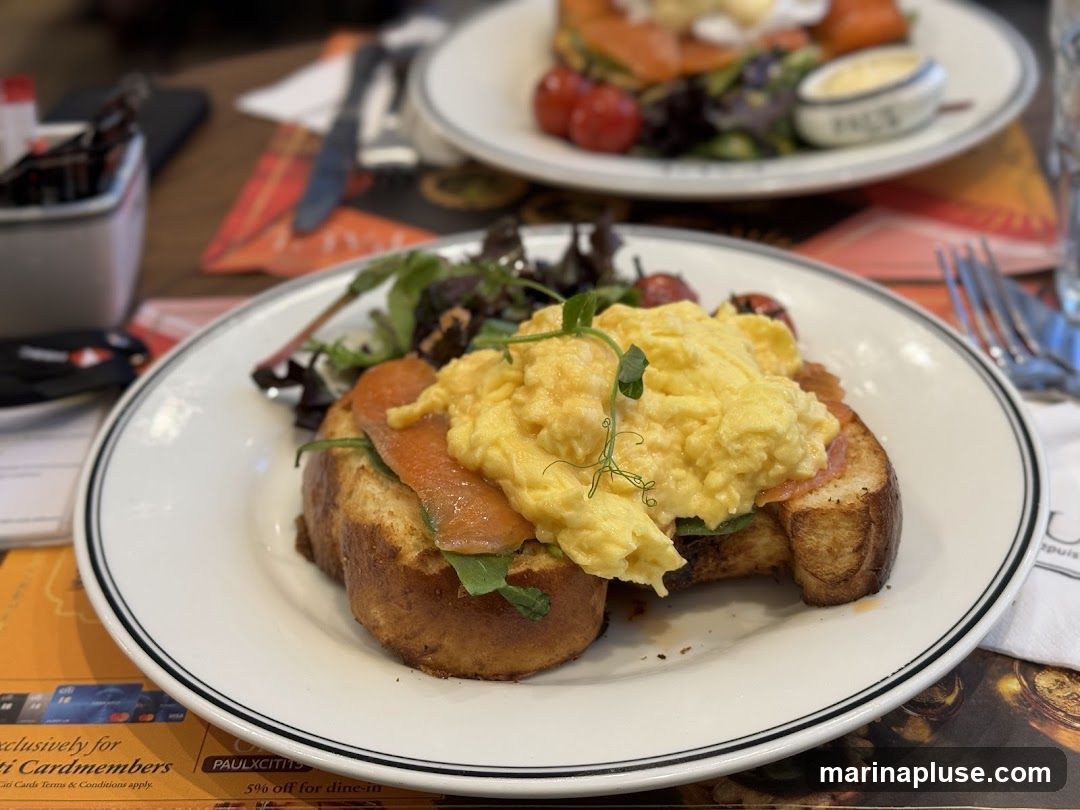 A plate of scrambled eggs and smoked salmon on toast, served with a side salad and cherry tomatoes. The dish is presented on a white plate with a black rim. In the background, another plate with a similar dish is visible. The table is set with cutlery and a napkin. A promotional flyer for Citi Cardmembers offering a 5% discount on dine-in is visible on the table.