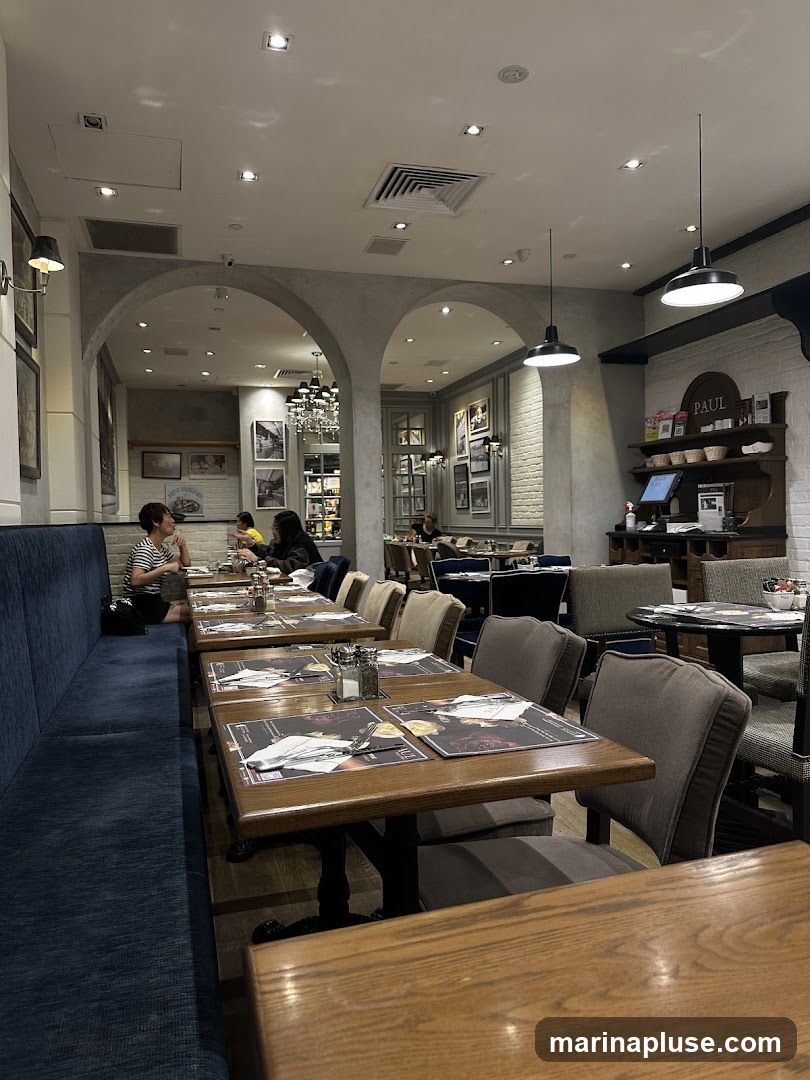 The image shows the interior of a restaurant with tables and chairs set up for dining. A long blue banquette runs along the left side of the room, with wooden tables and chairs facing it. In the background, more tables and chairs are visible, along with decorative elements like framed pictures and a chandelier. A shelf unit with the word "PAUL" is visible on the right side, with some products displayed on it.