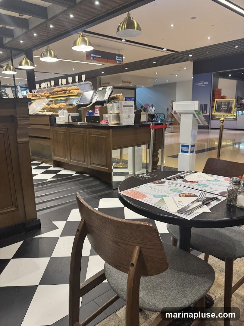 A view of a cafe interior with a display of pastries and baked goods behind a counter. A round table with chairs is set for dining, with cutlery and a menu visible. The floor has a black and white checkered pattern. In the background, there are signs for a car park and concierge, and escalators leading to other levels of the building. A sign for "PAUL" indicates the establishment is closed.