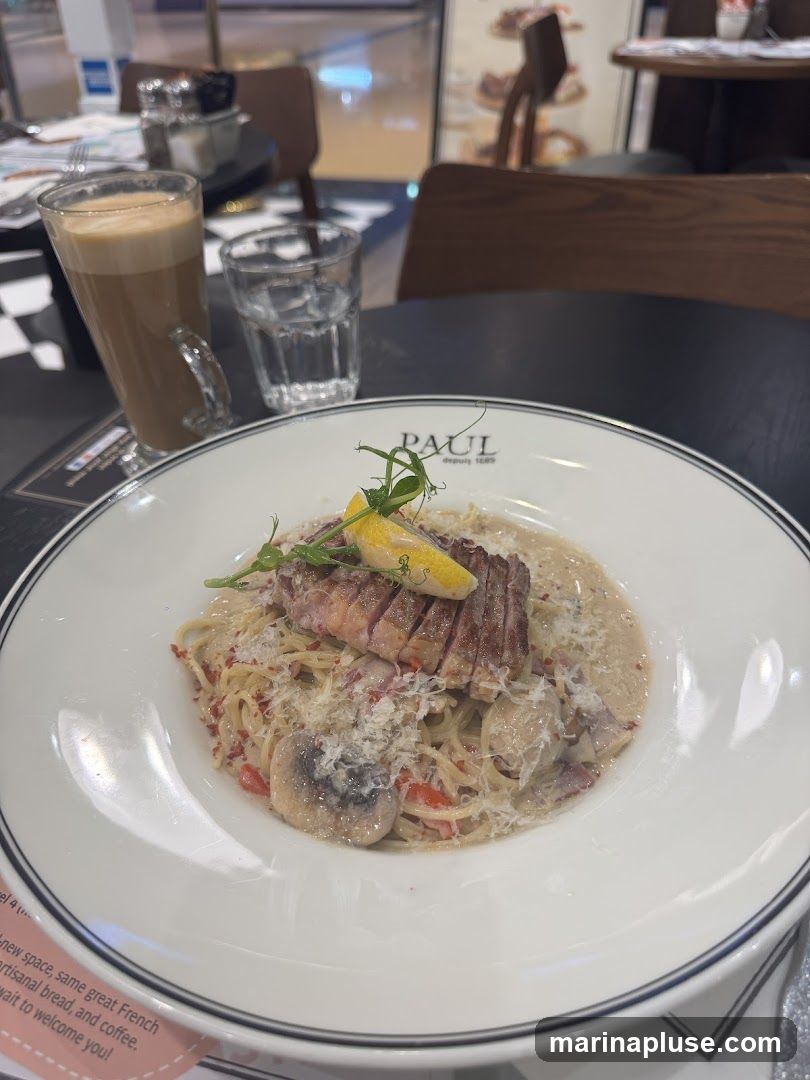 A plate of pasta with meat and mushrooms is served on a table with a glass of coffee and a glass of water. The plate has the PAUL logo on it. A pink flyer with text about a new space and French artisanal bread is visible at the bottom left.