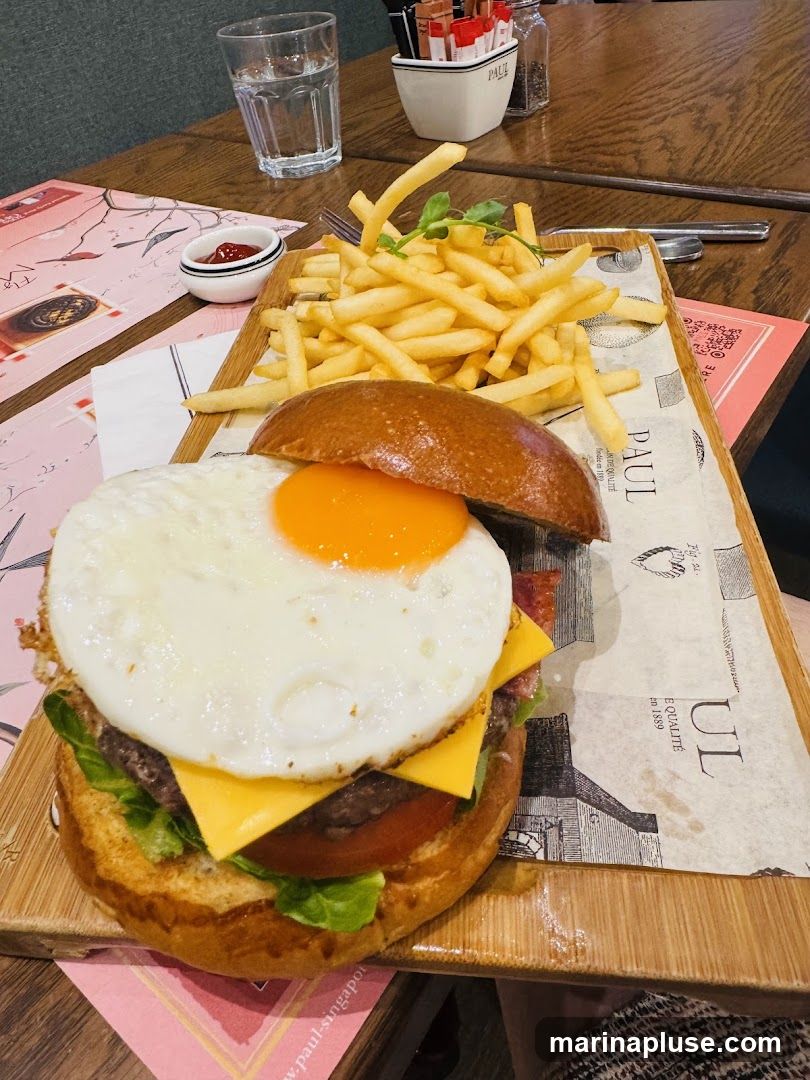 A close-up shot of a burger with a fried egg, cheese, lettuce, and tomato, served with a side of french fries and a small dish of ketchup. The meal is presented on a wooden board with branded paper. A glass of water and salt and pepper shakers are visible in the background.