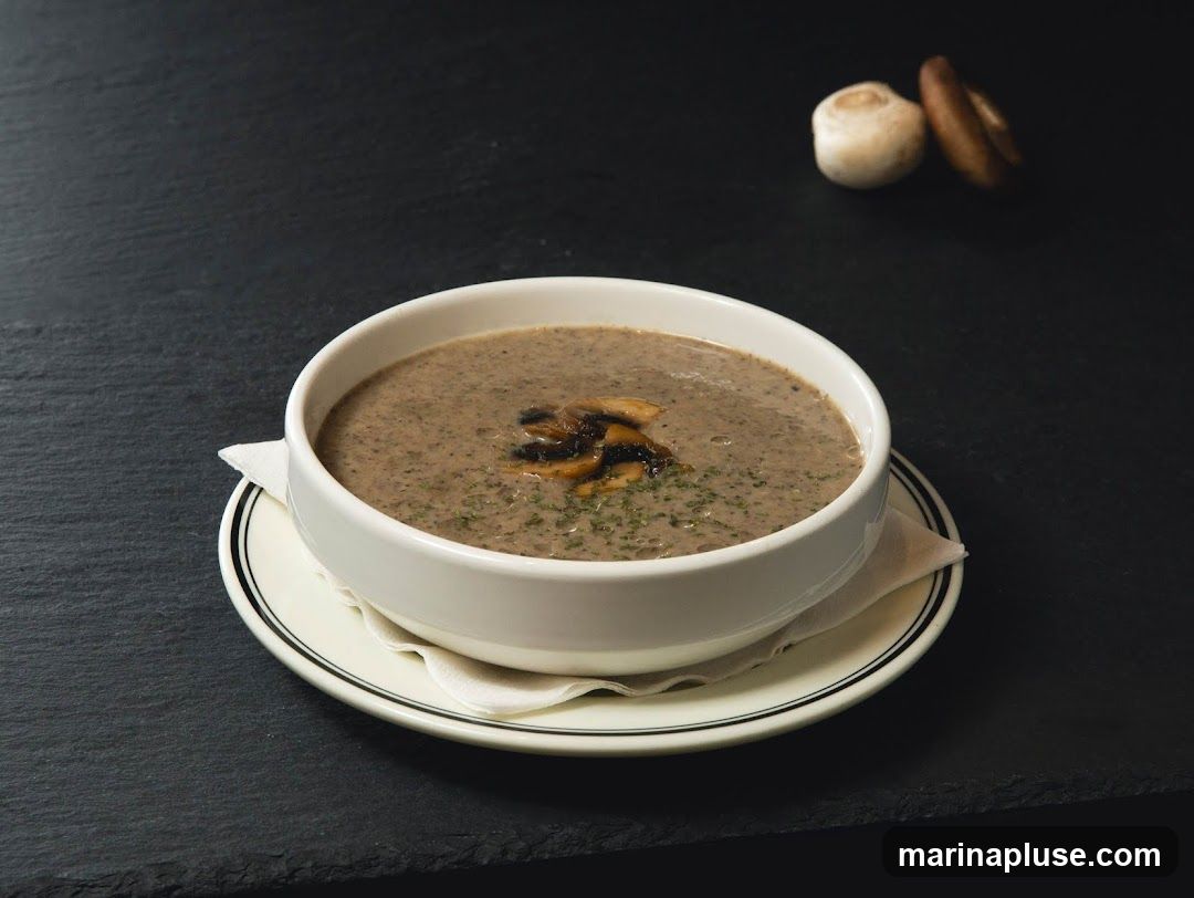 A bowl of creamy mushroom soup garnished with sliced mushrooms and herbs, served on a white plate with a black rim. Two whole mushrooms are visible in the background.