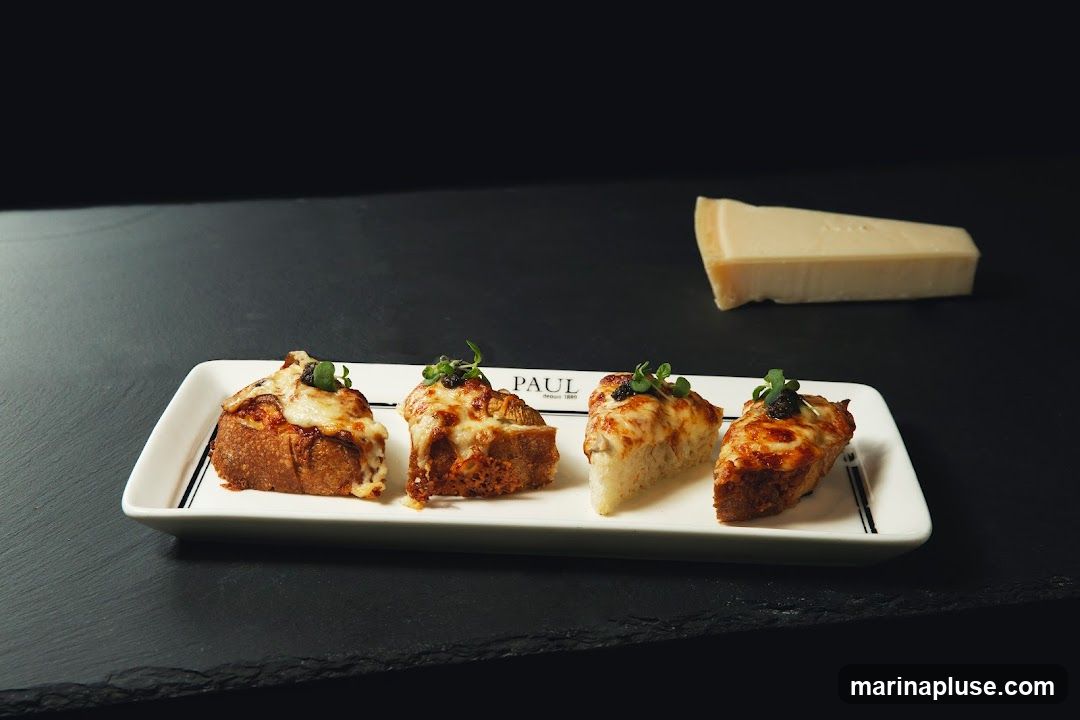 Four pieces of bruschetta, topped with melted cheese and microgreens, are arranged on a rectangular white plate with black trim. A wedge of hard cheese is visible in the background. The PAUL logo is printed on the plate.