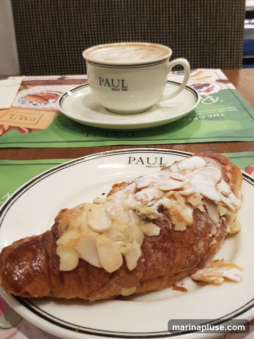 A close-up shot of an almond croissant on a white plate with a black rim, dusted with powdered sugar and topped with sliced almonds. Next to it, a white cup and saucer with the PAUL logo and "depuis 1889" printed on it, filled with a frothy coffee drink. The setting appears to be a cafe or bakery, with placemats and a textured background visible.