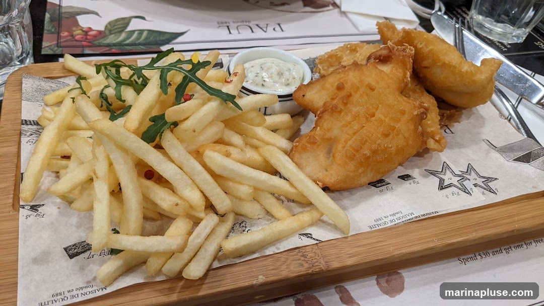A wooden board with a serving of fish and chips, a small bowl of sauce, and a garnish of arugula. The board is lined with paper featuring text and illustrations. A glass of water and cutlery are visible on the right side.