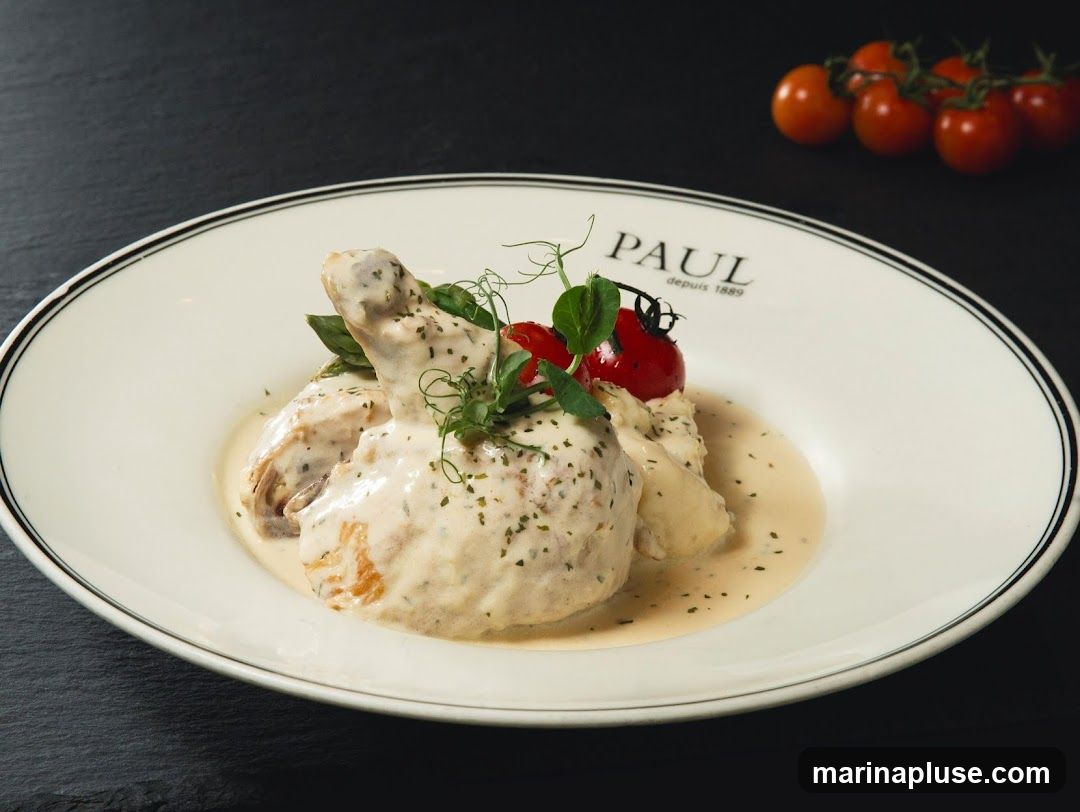 A close-up shot of a creamy chicken dish served on a white plate with a black rim. The plate has the PAUL logo and "depuis 1889" printed on it. The dish is garnished with microgreens and two cherry tomatoes. A bunch of cherry tomatoes is visible in the blurred background.