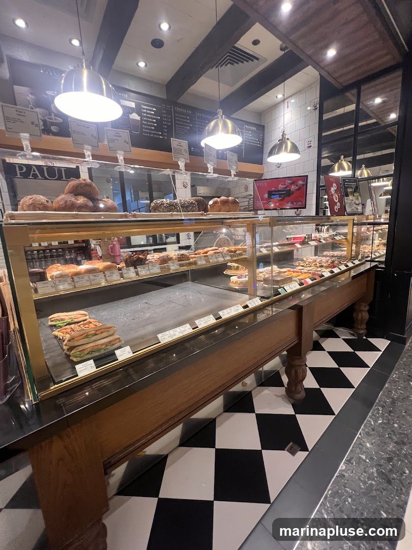 A well-lit interior view of a bakery display case filled with various baked goods, sandwiches, and pastries. The display features multiple shelves with breads, cakes, and other treats. The floor has a distinctive black and white checkered pattern. A prominent sign with the name "PAUL" is visible above the display.