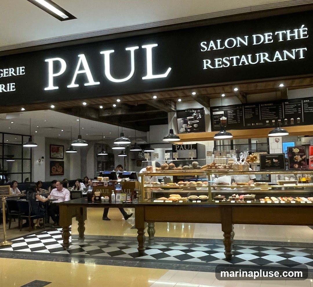 A view of the interior of a Paul bakery and restaurant, with a prominent sign displaying "PAUL" and "SALON DE THÉ RESTAURANT". The display cases are filled with pastries and bread. Customers are seated at tables in the background.