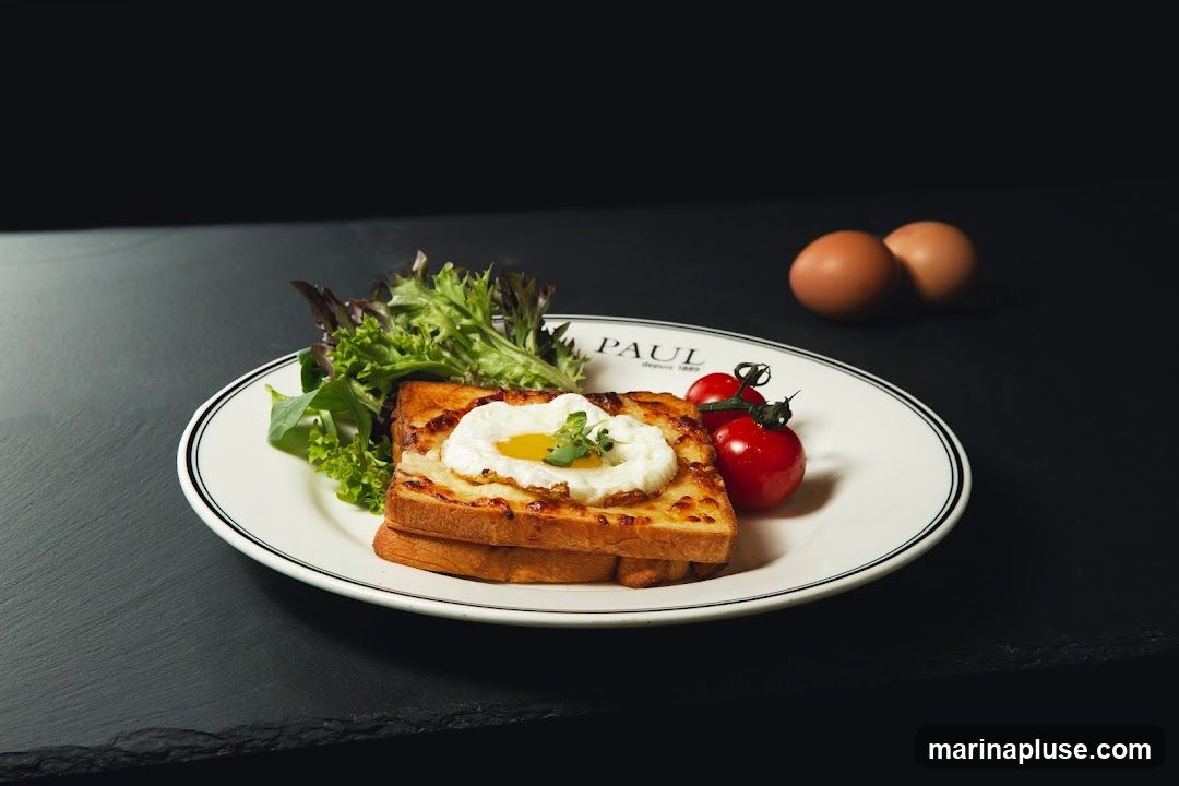 A Croque Madame sandwich topped with a fried egg and a sprig of parsley, served with a side salad and cherry tomatoes on a white plate with a black rim. The plate has the PAUL logo and "depuis 1889" printed on it. Two brown eggs are visible in the background.